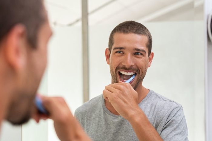 Smiling young man with toothbrush cleaning teeth and looking mirror in the bathroom oral health general dentistry dentist in Durham North Carolina